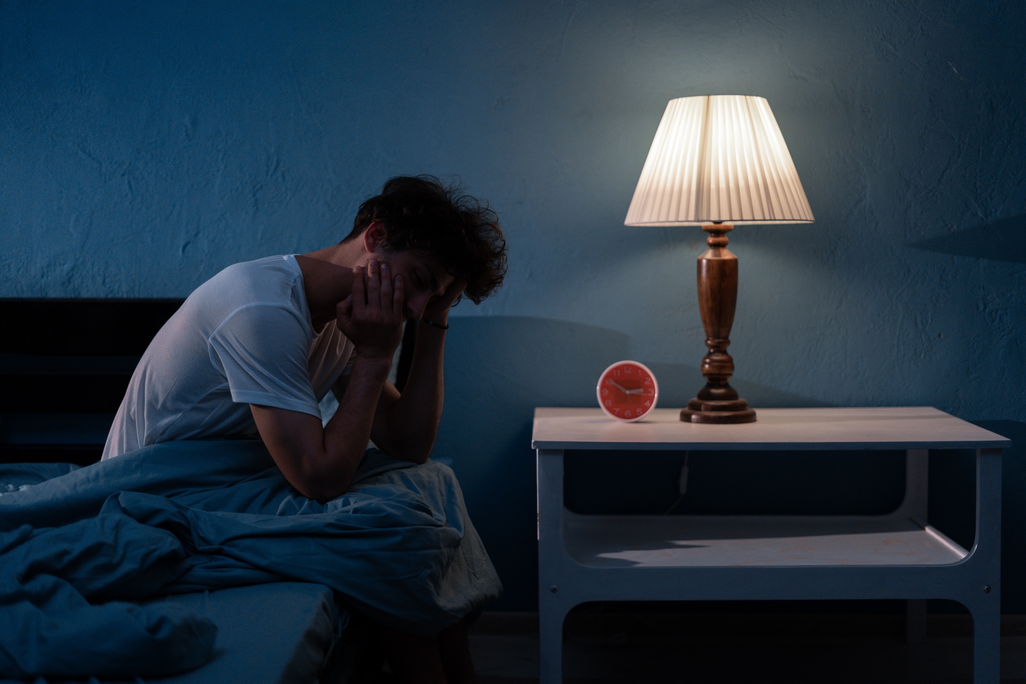 A tired man sitting on the edge of his bed at night, struggling with exhaustion and searching for sleep apnea near me to find relief from chronic fatigue and snoring.